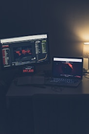 A dimly lit room with a desk featuring two screens displaying global COVID-19 statistics. A large monitor shows a world map with colored markers indicating various locations, alongside numerical data and graphs. A laptop is positioned nearby, also displaying a map with data, and an open notebook sits on the desk. A small lamp with a subtle glow provides additional lighting.