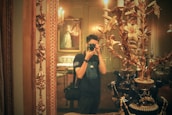A person is taking a photograph in an ornate room filled with vintage decor. This includes a large mirror with a decorative frame, an elaborate vase with golden flowers, and a classic painting in the background. The lighting is warm, creating a nostalgic atmosphere.