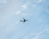 A commercial airplane is flying through a clear blue sky, offering a sense of movement and travel. The plane is seen from below, silhouetted against the vastness of the sky with subtle cloud patterns.