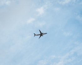 A commercial airplane is flying through a clear blue sky, offering a sense of movement and travel. The plane is seen from below, silhouetted against the vastness of the sky with subtle cloud patterns.