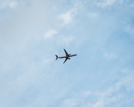 A commercial airplane is flying through a clear blue sky, offering a sense of movement and travel. The plane is seen from below, silhouetted against the vastness of the sky with subtle cloud patterns.
