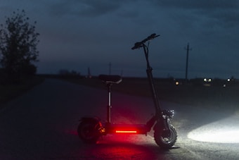 A black electric scooter is parked on a deserted road during twilight. It is illuminated by red lights on its platform, casting a faint glow on the ground. The background is dimly lit, with a few scattered lights from distant houses or streetlights visible. The setting appears to be rural, with a silhouette of a tree on the left side and power poles along the road.