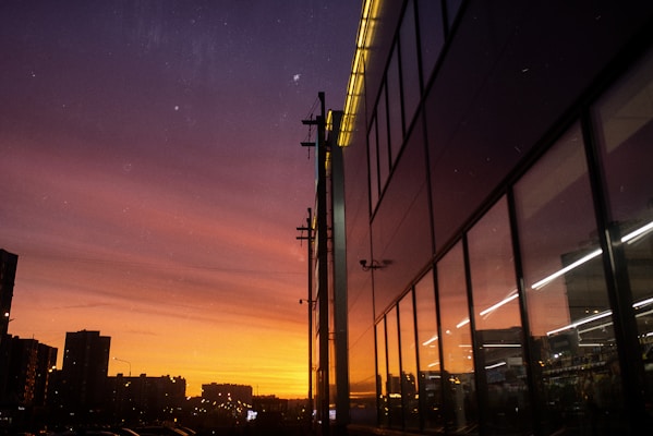A cityscape at sunset with a vibrant gradient sky transitioning from deep purple to orange. A modern glass building on the right reflects the sunset, while silhouetted buildings and power lines are visible on the left, creating a contrast with the bright sky.