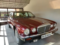 A classic burgundy car is parked indoors in a well-lit garage, showcasing a polished chrome grille and large round headlights. The interior appears to be beige, and a license plate is visible at the front.