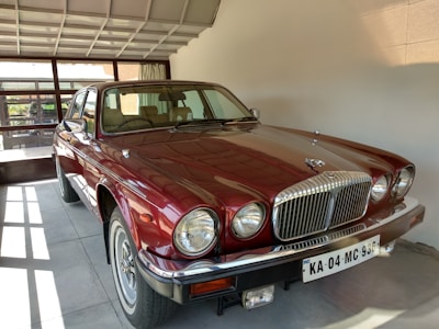 A classic burgundy car is parked indoors in a well-lit garage, showcasing a polished chrome grille and large round headlights. The interior appears to be beige, and a license plate is visible at the front.