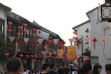 A bustling street market featuring traditional Chinese architecture adorned with numerous red lanterns and festive banners. People are walking along the street, which creates a lively and vibrant atmosphere.