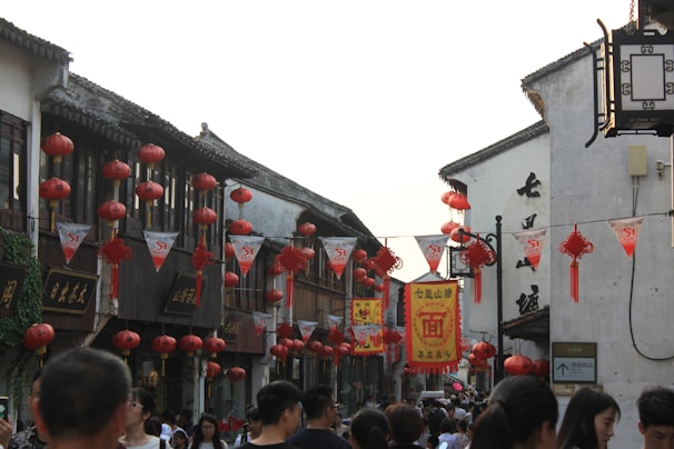 A bustling street market featuring traditional Chinese architecture adorned with numerous red lanterns and festive banners. People are walking along the street, which creates a lively and vibrant atmosphere.