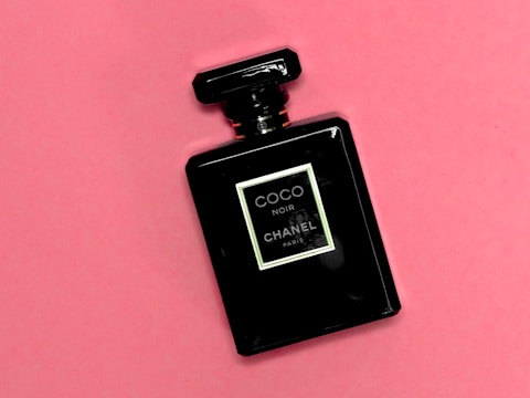 A sleek black perfume bottle with a square label and the text 'Coco Noir Chanel Paris' set against a soft pink background. The bottle has a classic, luxurious design.