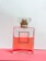 A glass perfume bottle filled with pink liquid rests on a pink surface. The bottle has a square shape and a minimalist label design with a clear cap.