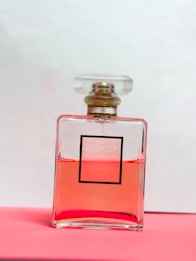 A glass perfume bottle filled with pink liquid rests on a pink surface. The bottle has a square shape and a minimalist label design with a clear cap.