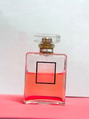 A glass perfume bottle filled with pink liquid rests on a pink surface. The bottle has a square shape and a minimalist label design with a clear cap.
