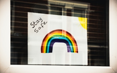 A colorful rainbow is painted on a sheet of paper with the words 'Stay Safe' written in black ink. In the top right corner, a yellow sun is depicted with rays extending outwards. The poster is taped to a window, and the room behind reflects on the glass.
