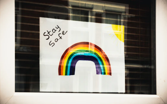 A colorful rainbow is painted on a sheet of paper with the words 'Stay Safe' written in black ink. In the top right corner, a yellow sun is depicted with rays extending outwards. The poster is taped to a window, and the room behind reflects on the glass.