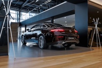A sleek black Mercedes-Benz E 200 coupe is parked in a modern showroom. The car's polished exterior reflects the ambient lighting. The interior of the showroom features wooden flooring, glass railings, and a contemporary design with metal and wood finishes.