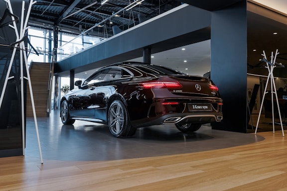 A sleek black Mercedes-Benz E 200 coupe is parked in a modern showroom. The car's polished exterior reflects the ambient lighting. The interior of the showroom features wooden flooring, glass railings, and a contemporary design with metal and wood finishes.