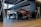 A sleek black Mercedes-Benz E 200 coupe is parked in a modern showroom. The car's polished exterior reflects the ambient lighting. The interior of the showroom features wooden flooring, glass railings, and a contemporary design with metal and wood finishes.
