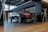 A sleek black Mercedes-Benz E 200 coupe is parked in a modern showroom. The car's polished exterior reflects the ambient lighting. The interior of the showroom features wooden flooring, glass railings, and a contemporary design with metal and wood finishes.