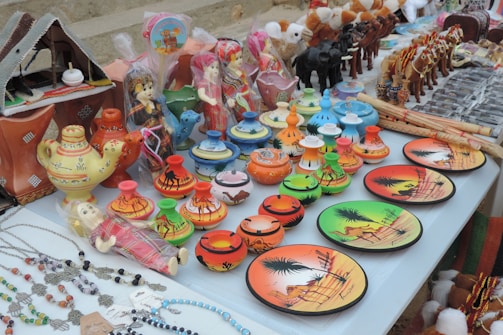 A display of colorful ceramic items and traditional souvenirs on a table. There are decorative plates with desert scenes, small pots with camel patterns, and miniature figurines of camels and people in vibrant attire. There are also crafted necklaces with beads and charms laid out to one side.