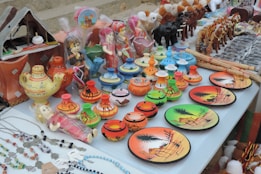 A display of colorful ceramic items and traditional souvenirs on a table. There are decorative plates with desert scenes, small pots with camel patterns, and miniature figurines of camels and people in vibrant attire. There are also crafted necklaces with beads and charms laid out to one side.