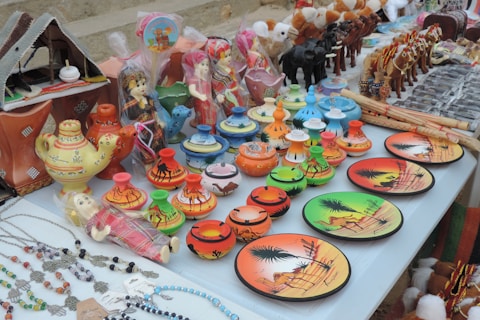 A display of colorful ceramic items and traditional souvenirs on a table. There are decorative plates with desert scenes, small pots with camel patterns, and miniature figurines of camels and people in vibrant attire. There are also crafted necklaces with beads and charms laid out to one side.