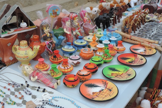 A display of colorful ceramic items and traditional souvenirs on a table. There are decorative plates with desert scenes, small pots with camel patterns, and miniature figurines of camels and people in vibrant attire. There are also crafted necklaces with beads and charms laid out to one side.