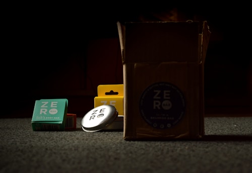 A collection of soap bars and a cardboard box are placed on a carpeted floor. The soap bars are packaged in colorful boxes, and one bar is visibly exposed, revealing a silver tin with the brand name on it. The items are partially illuminated, contrasting with the dark background.