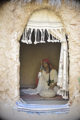 A woman is seated inside a small, rustic dwelling with walls made of earthen material. She is dressed in traditional clothing, adorned with colorful bracelets. Her expression appears contemplative as she rests her head on her hand. A stone grinding tool is placed in front of her on a fabric mat. The entrance to the dwelling is covered by a fringed cloth curtain, and a straw roof is visible at the top.