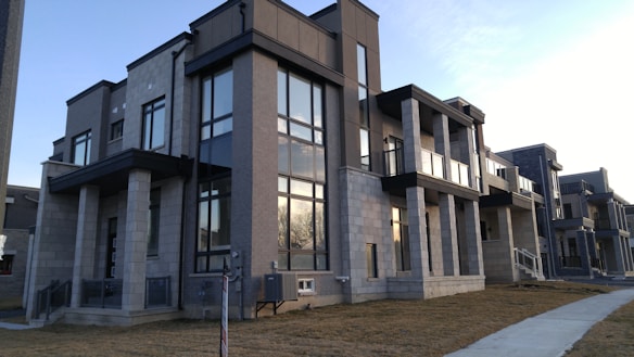 A modern apartment building with a symmetrical design featuring large windows and multiple balconies. The structure is constructed with light grey stone and dark trim, giving it a sleek and contemporary look. A small patch of grass and a concrete sidewalk can be seen in the foreground under a clear blue sky.