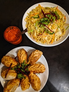 Two white plates are filled with different dishes. The top plate contains a serving of noodles mixed with vegetables such as carrots, cabbage, and bell peppers, garnished with cilantro. The bottom plate displays several fried dumplings arranged in a circular pattern, also garnished with herbs. A small glass filled with bright red sauce accompanied by a spoon is placed near the plates, set on a dark surface.