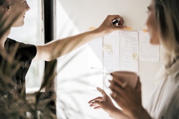 Two people are engaged in a collaborative activity, examining and discussing various notes and sketches pinned to a wall. One person is pointing with a pen, suggesting a brainstorming or planning session. The individuals are indoors, with soft lighting streaming through a window.