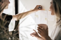 Two people are engaged in a collaborative activity, examining and discussing various notes and sketches pinned to a wall. One person is pointing with a pen, suggesting a brainstorming or planning session. The individuals are indoors, with soft lighting streaming through a window.