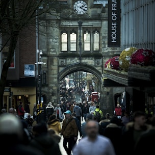 A bustling street scene in a historic city center, with a large crowd of people walking under an old stone archway adorned with a clock. Tall buildings and shops line the sides, including a visible House of Fraser sign and Joules store. Decorated spheres featuring star patterns hang prominently on the right side.
