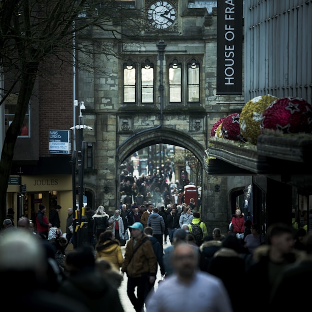 A bustling street scene in a historic city center, with a large crowd of people walking under an old stone archway adorned with a clock. Tall buildings and shops line the sides, including a visible House of Fraser sign and Joules store. Decorated spheres featuring star patterns hang prominently on the right side.