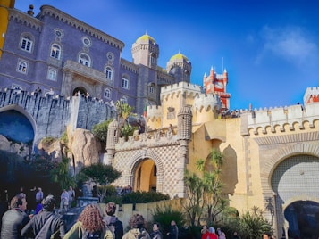 A vibrant and eclectic palace with various architectural styles, featuring bright yellow and red accents, intricate stonework, and lush greenery. The structure has towers with domes, and numerous tourists are gathered around the entrance.