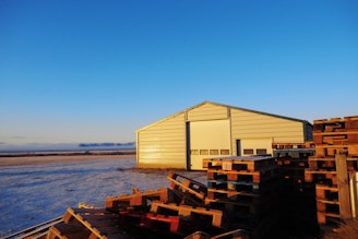 A large metal warehouse stands in a flat, open area, with a clear blue sky above. In front of the warehouse, there are stacks of wooden pallets. The ground is covered with a light dusting of snow, and the sunlight creates a warm glow on the building.