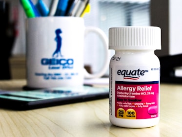 A white bottle of Equate Allergy Relief tablets is sitting on a wooden desk. The label is pink and blue, detailing the product as an antihistamine with 25 mg Diphenhydramine HCl. In the background, there is a white mug with pens of various colors, and a partially visible mobile phone.