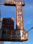 A rusted vintage sign with faintly visible words reading 'AUTO PARTS' and 'AUTO PAINTS', accompanied by some old light fixtures and electric wiring against a clear blue sky.