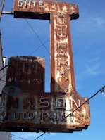 A rusted vintage sign with faintly visible words reading 'AUTO PARTS' and 'AUTO PAINTS', accompanied by some old light fixtures and electric wiring against a clear blue sky.