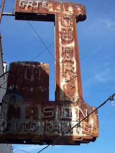 A rusted vintage sign with faintly visible words reading 'AUTO PARTS' and 'AUTO PAINTS', accompanied by some old light fixtures and electric wiring against a clear blue sky.