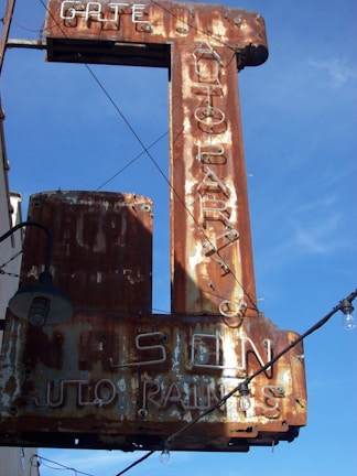 A rusted vintage sign with faintly visible words reading 'AUTO PARTS' and 'AUTO PAINTS', accompanied by some old light fixtures and electric wiring against a clear blue sky.