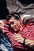 A person is resting on a bed, with sunlight creating shadow stripes across their face and body. They are wearing a red shirt and a black wristband, and there is a soft, textured blanket partially covering them.