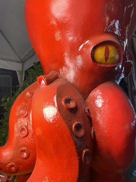 A close-up of a glossy, orange-red octopus sculpture with prominent suction cups and a large, vibrant yellow eye with a vertical slit pupil. The background includes hints of greenery and a tent-like structure.