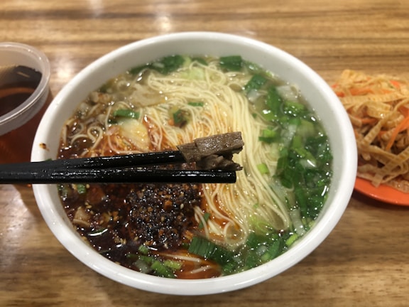 A steaming bowl of noodle soup contains finely chopped green onions, thin noodles, and a dark, savory broth. Black chopsticks hold a piece of meat over the bowl. On the side, an orange plate holds a serving of shredded vegetables, and a plastic cup is filled with a brown liquid, possibly tea. The setting suggests an informal dining environment with a wooden table.