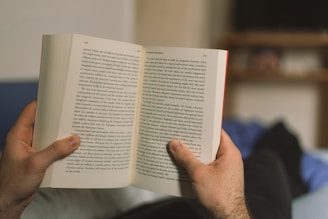 A person is holding an open book with both hands, comfortably reading while lying down. The book's pages are filled with text, suggesting an engaging read. In the background, a shelf with blurred items is visible, adding a cozy ambiance.