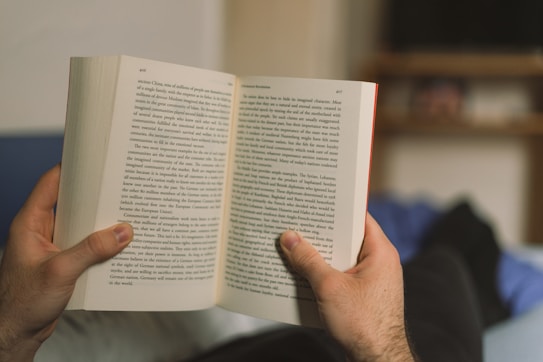 A person is holding an open book with both hands, comfortably reading while lying down. The book's pages are filled with text, suggesting an engaging read. In the background, a shelf with blurred items is visible, adding a cozy ambiance.