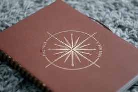 A close-up view of a brown notebook with a spiral binding, featuring a gold compass design on its cover along with the words 'Find Your True North'. The notebook is resting on a textured, grey fabric background.