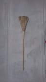 A traditional broom with a long wooden handle and straw bristles leaning against a plain gray wall. The broom is vertically aligned with its bristles at the top.