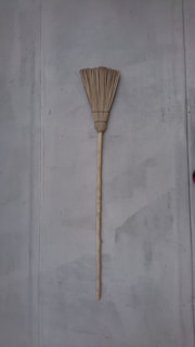 A traditional broom with a long wooden handle and straw bristles leaning against a plain gray wall. The broom is vertically aligned with its bristles at the top.