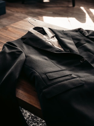 A dark suit jacket is neatly laid out on a wooden table with a white dress shirt underneath. The lighting casts a warm ambiance, emphasizing the texture of the fabric.