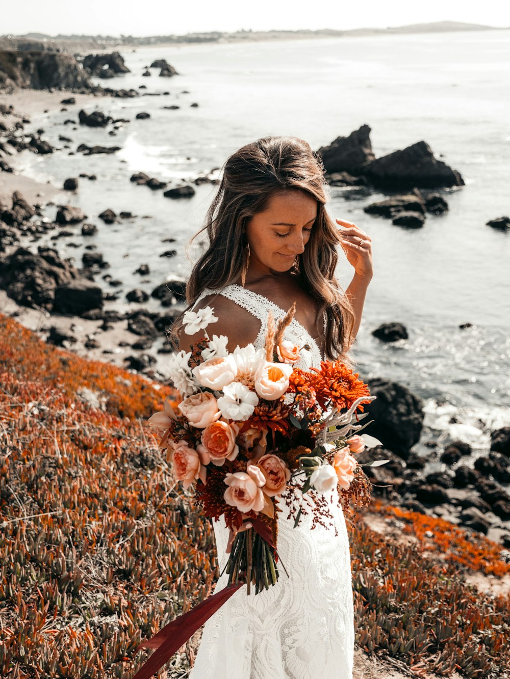 woman in white dress holding bouquet of flowers standing on rocky shore during daytime
